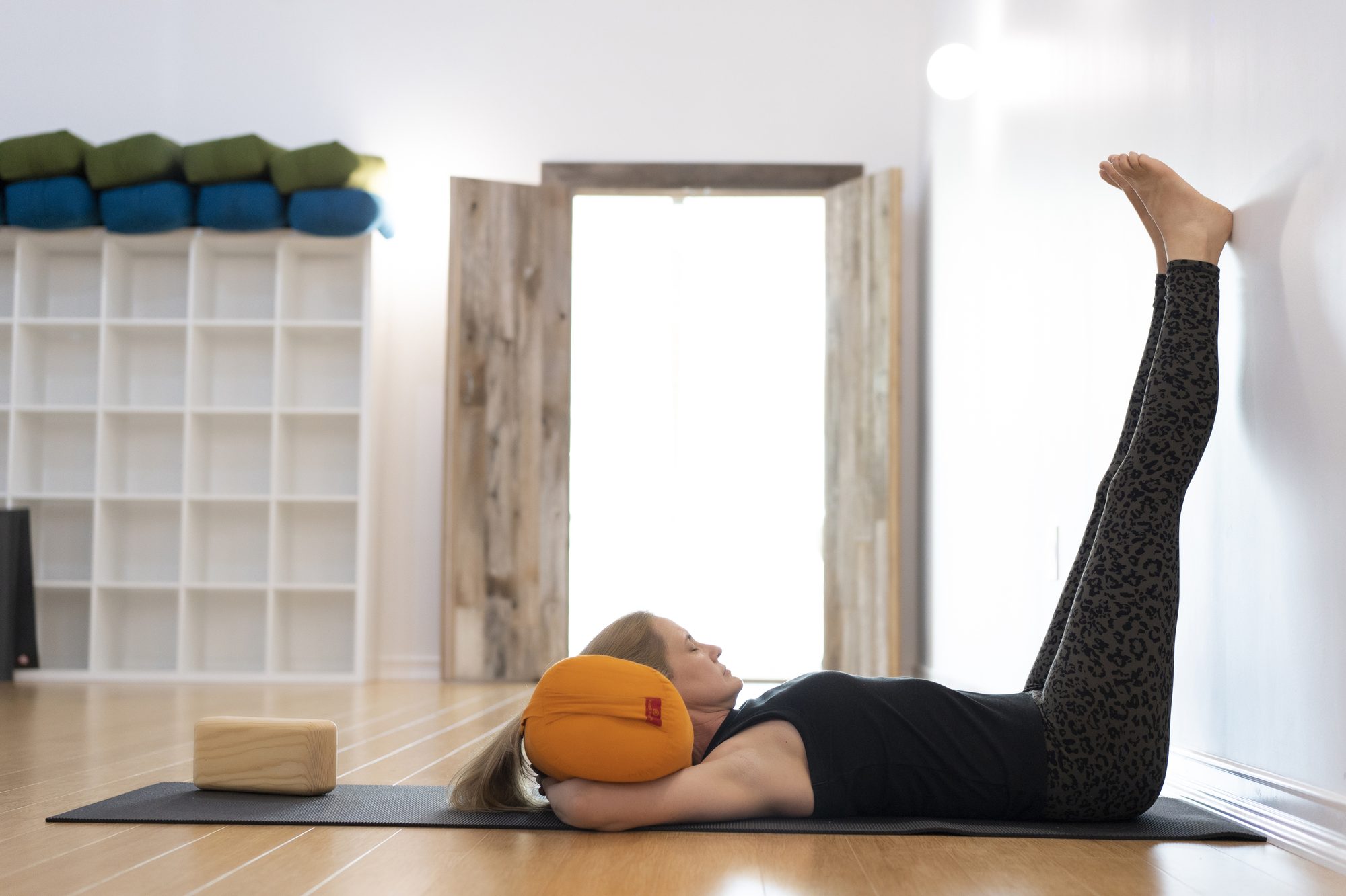 A student rests with both legs supported by the studio wall and an orange bolster beneath the head