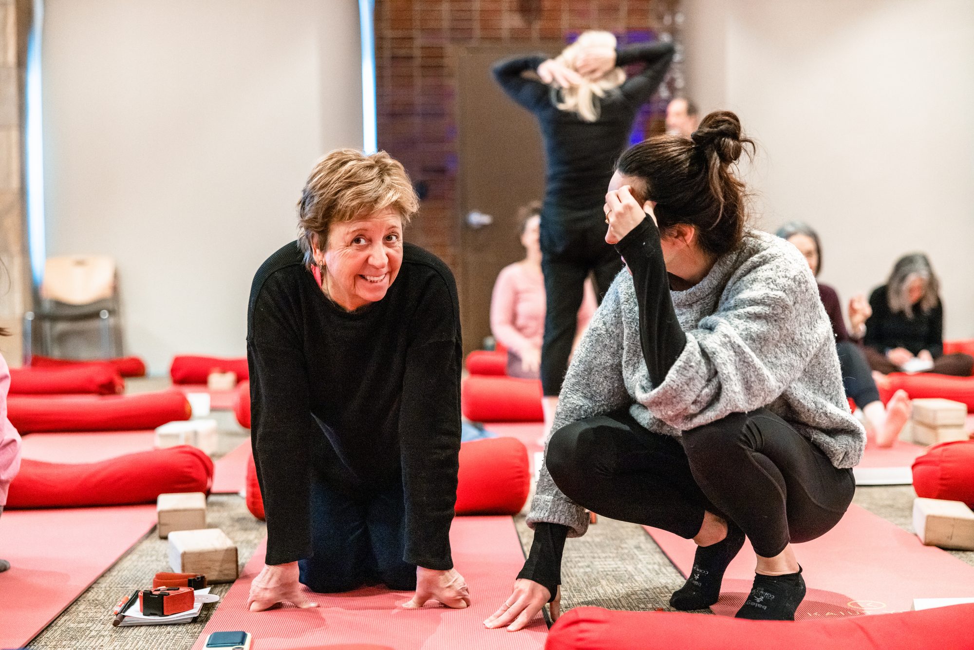 Renae crouched on the studio floor beside a student working on hands and knees, both mid-class on red mats